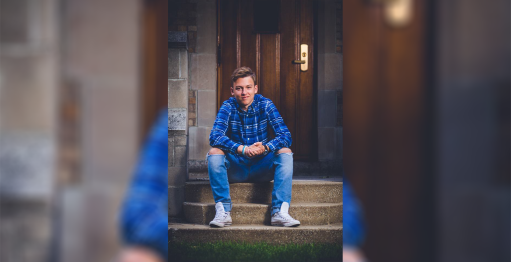 A person sits on stone steps in front of a wooden door. They wear jeans, a plaid shirt, and white sneakers.