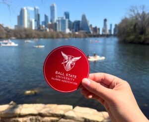 a hand holding Ball State Alumni Association coaster with White Beneficence in front of downtown Austin.